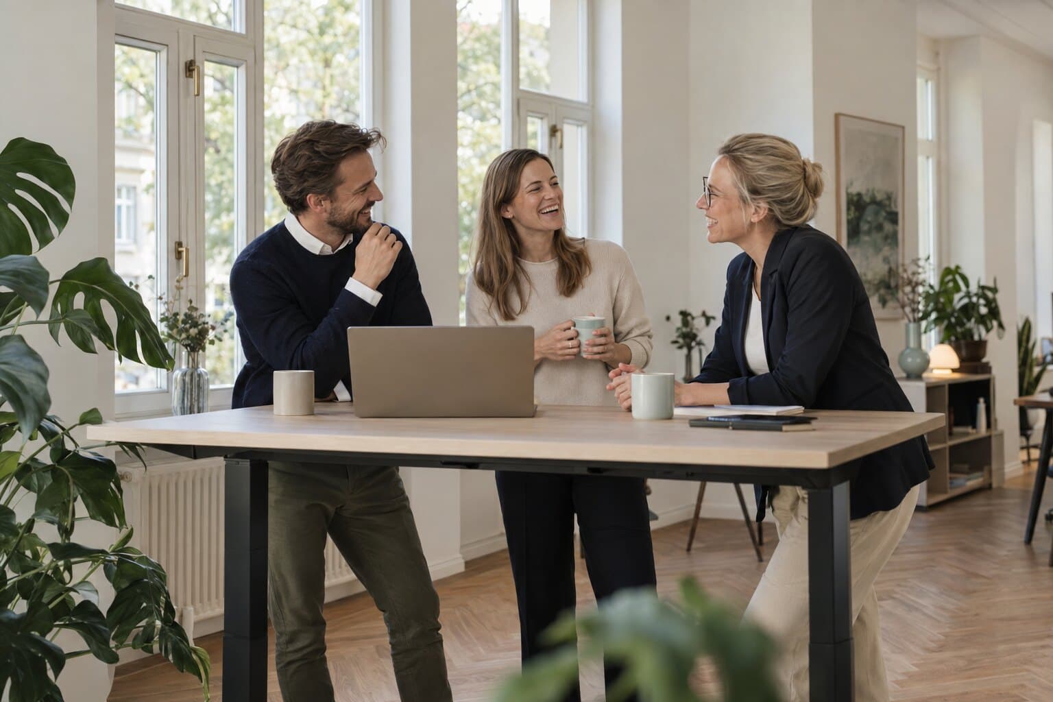 Three colleagues laughing together at a standing table with a laptop